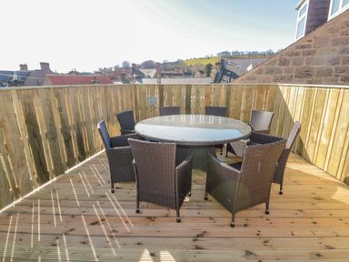 An outdoor wooden deck with a round glass table and eight wicker chairs at Cheviot Hills in Wooler