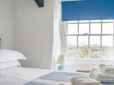 A bedroom with a bed with white linens and a blue striped blanket under a window with blue blinds at Cloam Cottage in Port Isaac