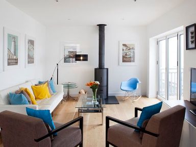 A living room with a white sofa and colorful pillows a glass coffee table a wood stove and a blue rocking chair at The Beach Hut in Polzeath