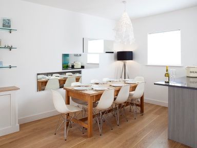 A dining area with a wooden table set for six and white chairs near a window at The Beach Hut in Polzeath