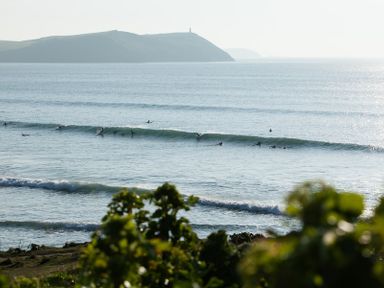 Surfers riding waves in the ocean with hills in the background at The Beach Hut in Polzeath