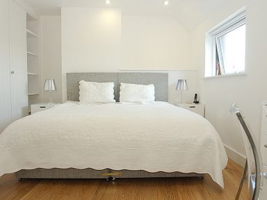 A bedroom with a large bed white bedding bedside tables with lamps an open window and wooden floor at The Beach Hut in Polzeath