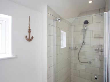 A shower area with a handheld shower head and tiled walls and a wooden anchor decoration on the adjacent white wall at The Beach Hut in Polzeath