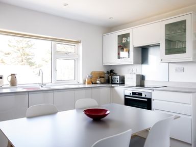A kitchen with white cabinets and appliances and a table with white chairs and a red bowl at Bella Vista in Polzeath
