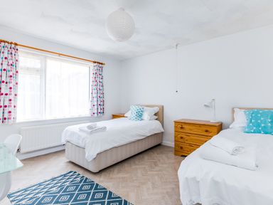 A bedroom with two single beds a window with patterned curtains a wooden dresser and a blue patterned rug at Bella Vista in Polzeath