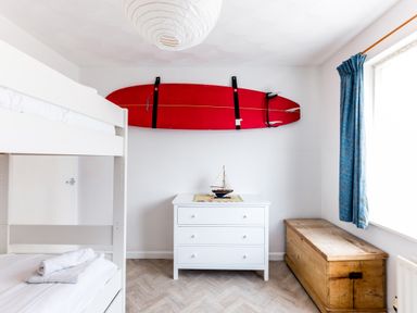 A bedroom with a bunk bed a white dresser with a model sailboat a wooden chest and a red surfboard mounted on the wall at Bella Vista Polzeath