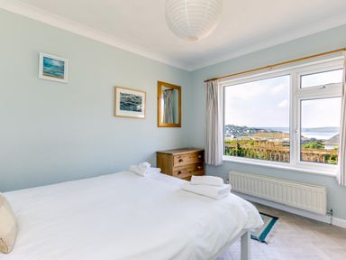 A bedroom with a bed with white linens towels on a wooden dresser a window with curtains and a view of a coastal town at Bella Vista in Polzeath