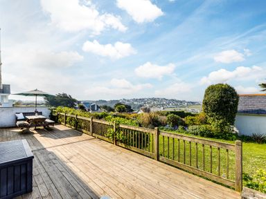 A wooden deck with a picnic table and umbrella overlooking a garden and distant houses at Bella Vista in Polzeath