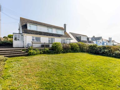 A grassy garden area with steps leading to a deck and a two-story house with multiple windows and a tiled roof at Bella Vista in Polzeath