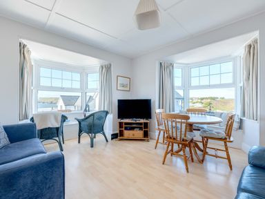 A living room with two blue armchairs by a window a wooden dining table with four chairs a TV on a wooden stand and two blue sofas at Pinewood Flat 5 in Polzeath