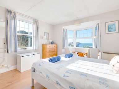 A bedroom with a bed with blue towels a wooden dresser a window with a sea view and two chairs at Pinewood Flat 5 in Polzeath