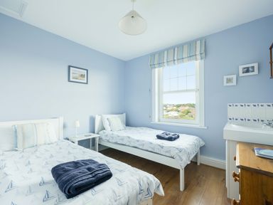 A bedroom with two single beds with sailboat patterned bedding a small bedside table a window with striped blinds and a washbasin with cabinetry at Pinewood Flat 5 Polzeath