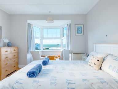 A bedroom with a bed, two blue rolled towels, two chairs near a bay window overlooking the sea at Pinewood Flat 5 Polzeath