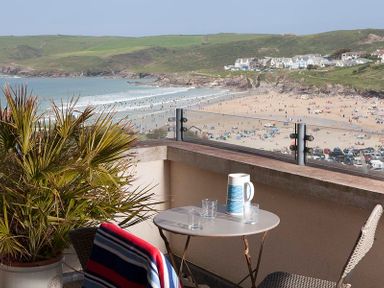 A balcony with a table and chairs overlooking a beach with people and houses on hills at Kellan in Polzeath