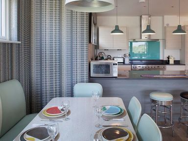 A dining area with a set table and light blue chairs next to a kitchen with appliances and pendant lights at Kellan Polzeath