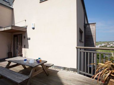 An outdoor wooden picnic table with mugs and a teapot on a deck next to a beige building with a small covered entrance at Kellan in Polzeath