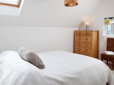 A bedroom with a bed a wooden chest of drawers a lamp a wicker chair and a skylight window at Kellan in Polzeath