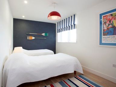 A bedroom with two single beds a striped rug a navy accent wall with two oars a striped window blind and a framed poster at Kellan Polzeath