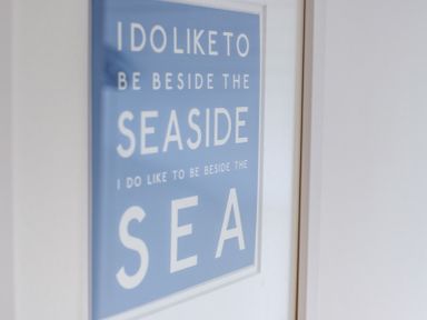 A framed blue and white sign on a wall with the text I do like to be beside the seaside at The Moorings in Rock