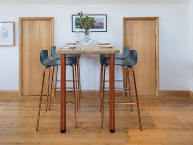 A dining area with a wooden table and four high chairs with blue seats two wooden doors and framed pictures on the wall at Sandy Hills 8 in Rock