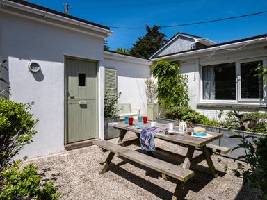 An outdoor patio area with a wooden picnic table set with cups plates and a hat beside a white building with a green door and window at St Michaels in Rock