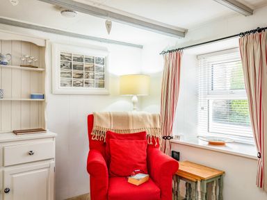A red armchair with cushions and a throw next to a window with striped curtains and a small wooden table at St Michaels in Rock