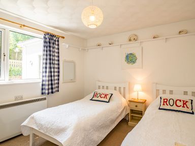 A bedroom with two single beds with white bedspreads and pillows that say rock a nightstand with a lamp between the beds a window with checkered curtains and a wall shelf with decorative plates at St Michaels in Rock