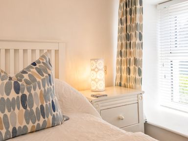 A bedroom corner with a white bed and patterned pillows next to a white bedside table with a decorative lamp and patterned curtains at St Michaels in Rock