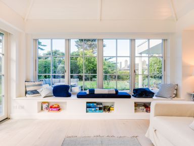 A seating area with cushions and board games under large windows overlooking a garden at Greystones in Daymer Bay