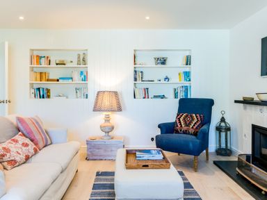A living room with a white sofa, a blue armchair, built-in bookshelves, a lamp on a wooden side table, and a fireplace at Greystones in Daymer Bay