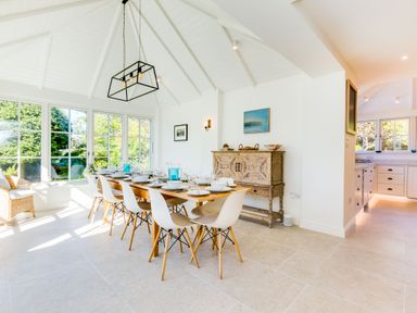 A dining room with a long wooden table set with white bowls and glasses and white chairs near large windows and a wooden cabinet at Greystones Daymer Bay