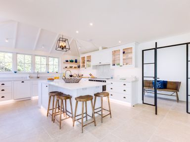 A kitchen with a white island and four wooden stools glass cabinets and a bench behind glass doors at Greystones in Daymer Bay