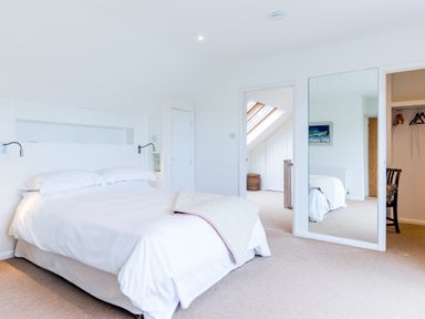 A bedroom with a double bed white bedding built in shelves wall lamps a large mirror and an open closet door at Greystones in Daymer Bay