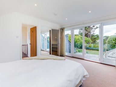 A bedroom with a bed, wooden door, mirror door, glass doors leading to a balcony, and a view of trees outside at Greystones in Daymer Bay