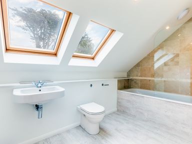 A bathroom with two skylight windows a wall-mounted sink a toilet and a bathtub with a glass screen at Greystones in Daymer Bay