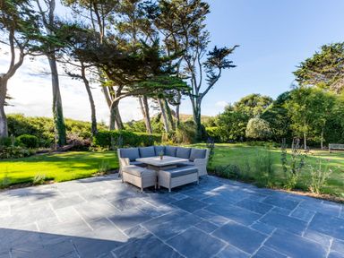 An outdoor patio with gray stone tiles and a corner sofa set in front of a grassy garden with trees and shrubs at Greystones in Daymer Bay
