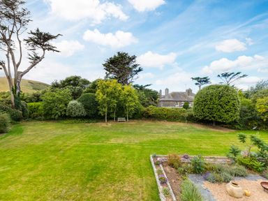 A garden with a green lawn trees shrubs a stone house in the background and a small patio with plants