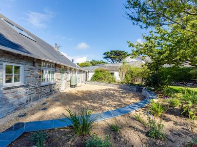 A stone house with a gravel courtyard and garden beds with plants and trees at Greystones in Daymer Bay