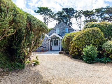 A gravel driveway leading to a stone house with a white door and two windows surrounded by tall trimmed hedges and trees at Greystones in Daymer Bay