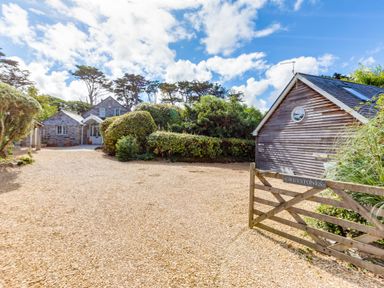 A gravel driveway with a wooden gate leading to a stone house and a wooden building surrounded by bushes and trees at Greystones in Daymer Bay
