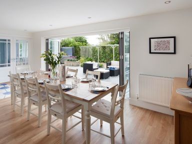 A dining room with a wooden table and white chairs set for a meal next to an open patio door leading to a garden with outdoor seating at Cherrytrees in Rock