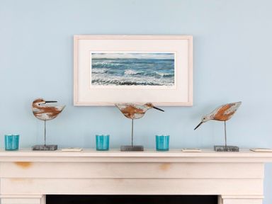 A fireplace mantel with three wooden bird sculptures blue candle holders and a seascape painting on a blue wall at Cherrytrees in Rock