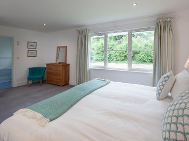 A bedroom with a bed, teal chair, wooden dresser with mirror, and large window showing greenery at Cherrytrees in Rock