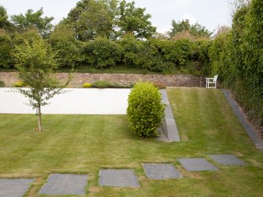 A garden with neatly trimmed grass a small tree a bush stone slabs forming a path and a white chair near a stone wall at Cherrytrees in Rock