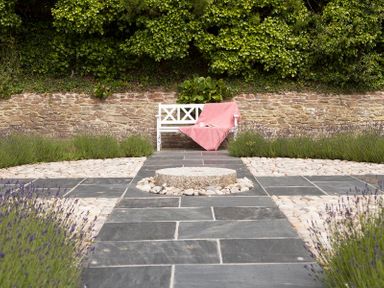 An outdoor garden with a white bench draped with a pink cloth surrounded by lavender plants and stone paving at Cherrytrees in Rock