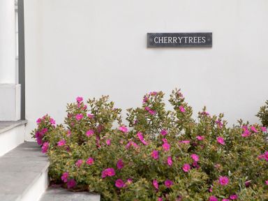 A white wall with a sign reading CHERRYTREES above a bed of pink flowering plants at Cherrytrees in Rock
