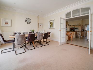 A dining room with a long table, chairs, wall clock, paintings, and an open door leading to a kitchen at The Haven in Daymer Bay