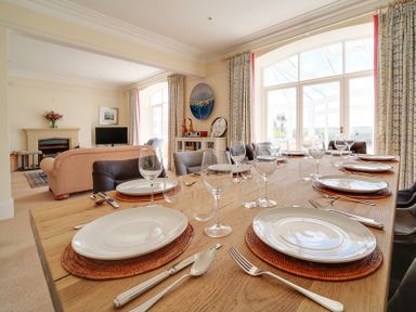 A dining table set with plates glassware and cutlery in a room with sofas a fireplace and large windows at The Haven in Daymer Bay