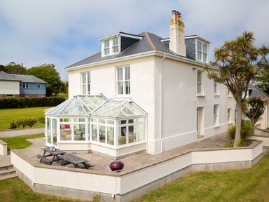 Exterior of a white house with a glass conservatory and patio with picnic benches at The Haven in Daymer Bay