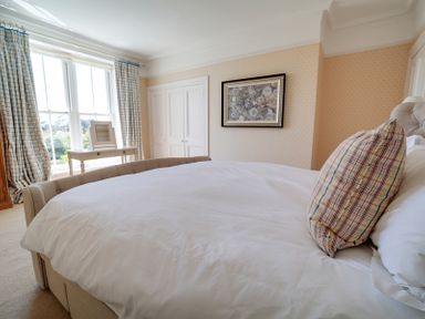 A bedroom with a large bed white bedding a plaid pillow a window with curtains a table and a framed picture on the wall at The Haven in Daymer Bay